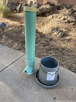 Photo of two chicken feed containers placed outdoors on cement and dirt ground with rocks in background. One is tall green plastic vertical feeder and the other is galvanized metal bucket feeder with base.