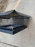 Angle view of the black metal chimney cap showing diamond-pattern mesh sides, rectangular base, and flared top hood placed on concrete.