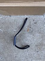 Single black metal coat hook laying on concrete showing shape and size.