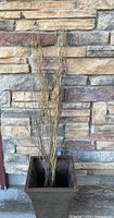Photo showing the tall square copper-colored metal container filled with assorted decorative twigs against a stone wall.