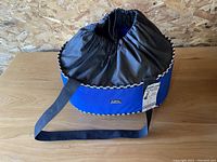 Blue Katie's Bumpers tote with black drawstring top and shoulder strap on wooden table.