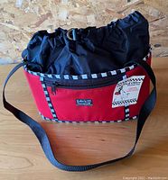 Full view of the red tote with black and white trim, adjustable black shoulder strap, and brand label on front.