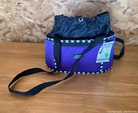 Front and side view of the purple Katie's Bumpers Oval Office bag showing the black and white checkered trim, black strap, and front label.