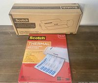 Scotch Thermal Laminator in its original box and a box of Scotch Thermal Laminating Pouches on a wooden table.