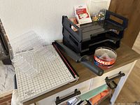 Photo showing black 3-tier document tray, clear acrylic file sorter, guillotine paper cutter, and tape dispenser with Staples tape on a brown table