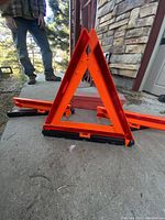 Side angle view of one folded orange triangle road hazard sign showing its shape and black base, placed on concrete near a building.