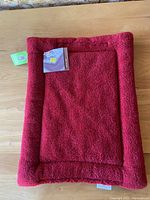 Full view of the rectangular red pet bed with raised edges on a wooden surface with attached tags.