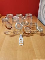 Eight sealable glass jars of various sizes and brands with metal clamp lids and seals arranged on table with a jar lid opener tool in front.