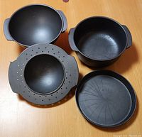 Four black non-stick Betty Crocker pans arranged on a wooden surface showing the flat round pan, the two deeper pans (one with holes), and the smaller insert pan