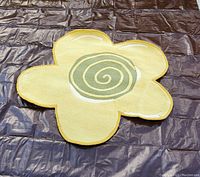 Full view of the yellow flower-shaped rug with a green and beige spiral center, placed on a dark surface.