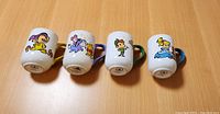 Four white ceramic Disney mugs displayed with character artwork side up showing Mickey Mouse, Dumbo, Pinocchio, and a blue-haired fairy character each with colorful designs and matching handle colors.