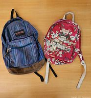 Top-down view of two adult backpacks on a wooden surface, left Jansport in blue stripes, right Cath Kidston in red floral pattern