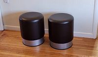 Two round dark espresso leather bar stools with chrome base, placed side by side on wooden floor against a white wall.