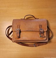 The front outer appearance of the brown leather satchel showing dual buckle closures and top handle with shoulder strap.