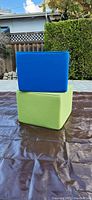 Side view of two cube-shaped outdoor poufs stacked, bright blue on top of lime green, set on a brown tarp outdoors with some backyard background.