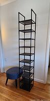 Vertical shoe rack with multiple shelves and round upholstered stool placed side by side on wooden floor against white wall.