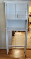 Front view of the white bathroom cabinet showing two doors and legs designed to fit over a toilet.