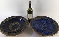 Two large pottery plates side by side with a bottle for scale, showing size and design details.