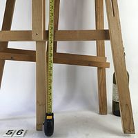 Close-up photo of the bottom portion of a wooden easel with measuring tape showing about 56 inches tall. Includes part of a wine bottle for scale.