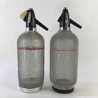Two vintage glass seltzer bottles with metal mesh covers and black dispensing heads, one bottle missing part of the dispenser as shown