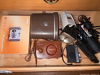 Full lot photo showing all items in a drawer: Argus camera in leather case, Canon PowerShot box and case, Sylvania flash, GE transistor radio, leather binoculars case