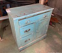 Wide shot of the blue wooden cabinet showing front and side with drawers and door, surface chipped and scratched.