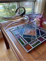 Photo showing the clear glass swan, purple bowl, pink blown glass piece, and stained glass window hanging on a wood table near window.
