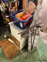 Overall view of laundry items including wicker hamper, blue plastic basket with trays, ironing board, drying frame, and a Magic Duster handle.