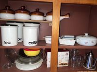 Photo of white ceramic kitchen canisters labeled tea, flour, and coffee with white lids, along with ceramic soup bowls with brown lids on upper shelf, glass pie plates, and various kitchenware below
