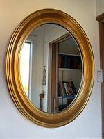 Full view of oval mirror hung on wall showing gold toned frame and reflection of bookshelf.