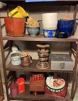 Shelving unit displaying multiple ceramic pots, a wooden candle holder, metal lockbox without key, and vintage tins. Pots vary in color, size, and design, some stacked inside others. Candle holder is centrally placed on middle shelf. Metal lockbox is silver and rectangular.