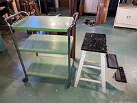 Vintage green three-tier rolling cart and white wooden retractable step stool shown together on basement floor.