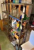 Side view of two metal shelving units showing five shelves each, filled with various household items such as glass bottles, cleaning products, and boxes. Shelves are scratched with visible signs of use.