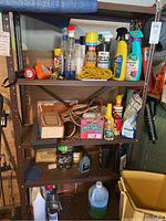Brown metal shelving unit with multiple shelves containing various garage and household items including chemicals, oils, cords, and small tools; rust is visible on the metal frame.