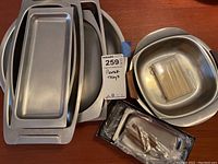 Multiple rectangular and oval stainless steel trays stacked and arranged, alongside a round bowl and a boxed set of stainless steel serving forks.