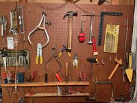 Photo of various hand tools mounted on pegboard including calipers, pliers, screwdrivers, rulers, hammer, saw, and drill bits.