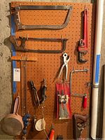 Pegboard wall with large metal saws, pipe wrench, leather punch, pliers, and hanging tools including a red handled tool and metal scrapers.