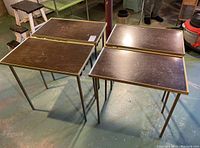 Four vintage folding tray tables arranged on basement floor. Shows wood grain tabletops with metal edging and folding metal legs.