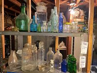 Shelf of assorted vintage glass bottles and jars including green, clear, and blue colors with some corked and others with lids, displayed on metal shelving in a basement setting.