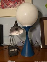 Two vintage lamps on desk: a blue mid-century modern lamp with large white glass globe shade showing paint chipping; next to an adjustable arm lamp with black metal shade and heavy transformer base.