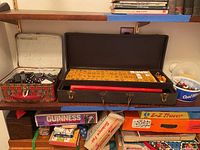 Photo shows vintage mahjong set tiles arranged in case, vintage boxed dominoes in lunchbox on shelf.