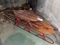Two vintage sleds stacked together against a basement wall, showing wooden seats and red metal frames