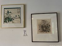 Two framed prints hanging on a white wall. Left print features Asian theme with birds and flowers, right print is black and white signed Anne Eldridge print with plants and flamingo.