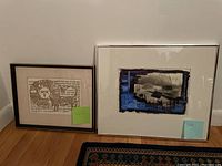 Photo showing two framed pieces of wall art side by side on floor, one abstract with blue and black, one stylized lion with Hebrew text.