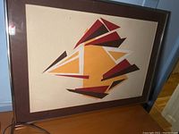 Full view of one framed art piece showing sharp angled geometric shapes in orange, red, brown, and white on textured cream background with brown mat and silver frame.