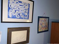 Three framed pieces of wall art hanging on a blue wall: top left is blue geometric maze print, bottom left is an embossed white floral piece, right is an abstract colorful piece in blue frame hung with pink string
