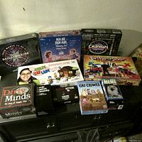 Photo showing 11 adult party games on a black cabinet surface against a plain wall. Games include well-known party and humorous games aimed at adults.