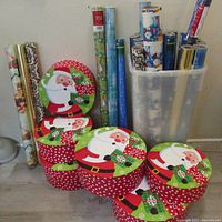 6 round Christmas gift boxes with Santa design, arranged on floor alongside standing rolls of gift wrap.