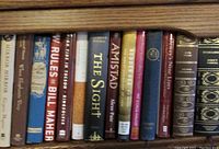 Photo of one shelf of hardback books including titles like 'New Rules' by Bill Maher, 'The Sight', 'Amistad', 'Mirror Mirror', and 'When Elephants Weep'