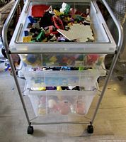 Full view of three-tier metal rack with four clear plastic bins containing LEGO bricks on top and vintage kids toys on the bottom two bins.